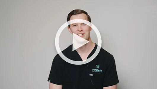 Dentist in black scrubs smiling during video interview with play button overlay